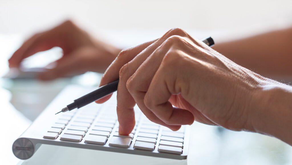 Close-up of hands typing on computer keyboard while holding a pen.
