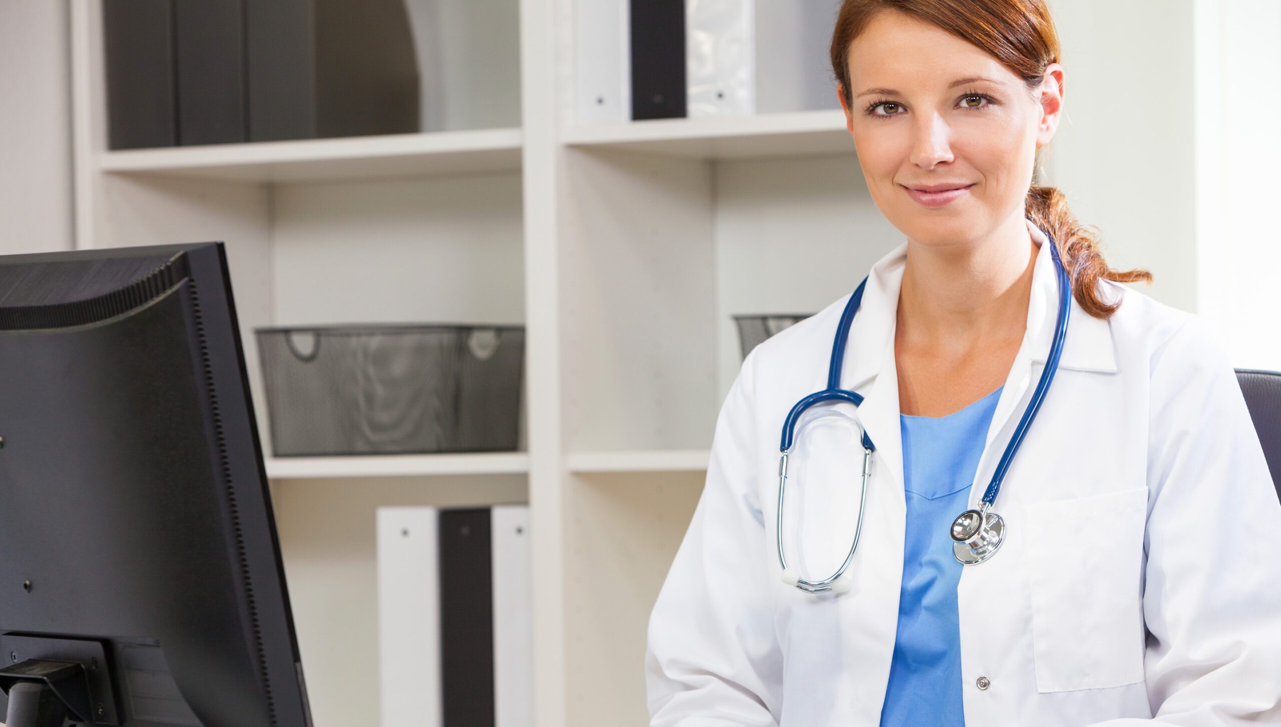 Female doctor smiling at desk with stethoscope in medical office.