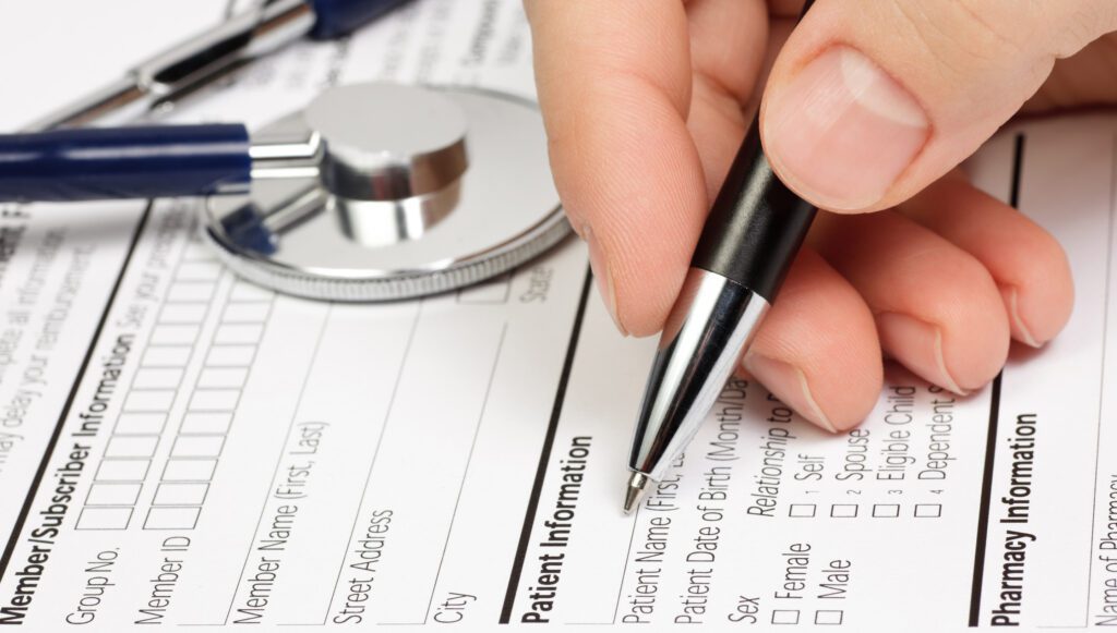 Close-up of hand filling out patient information form with stethoscope nearby.