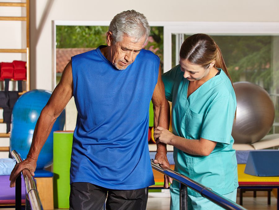 Physiotherapist supporting an older man as he practises walking between parallel bars during a rehab session.