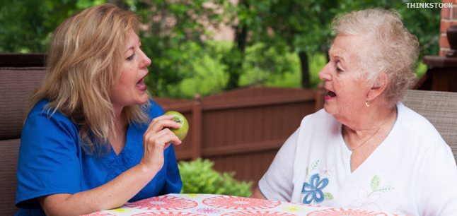Caregiver in blue scrubs engaging in a speech exercise with an elderly woman outdoors.