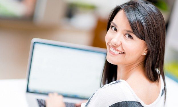 Person typing on a laptop and smiling back toward the viewer in a bright indoor setting.