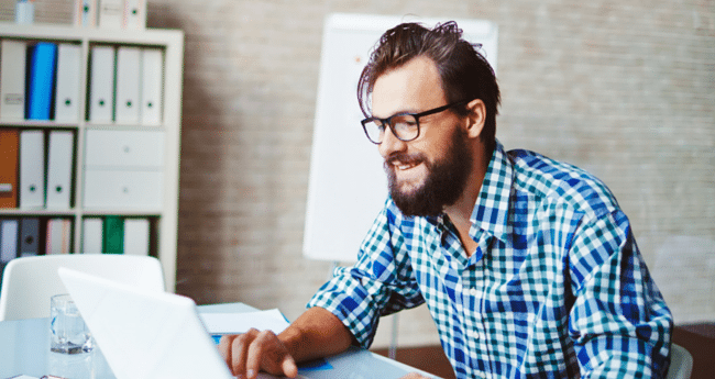 Smiling man with glasses working on a laptop in a modern office setting.