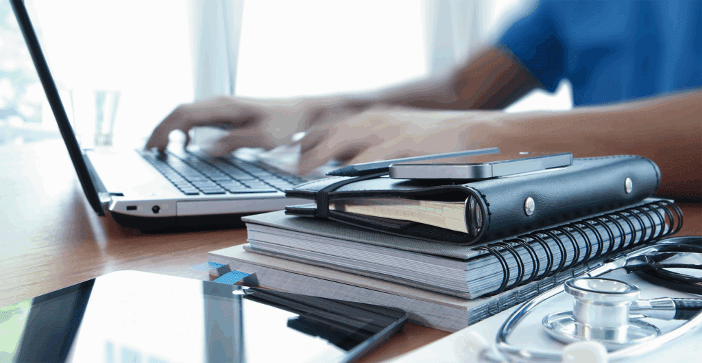 Healthcare professional working on a laptop beside stacked notebooks, a phone and medical tools including a stethoscope.