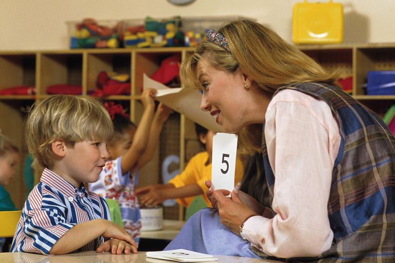 Teacher holding up a flashcard with the number 5 while interacting with a young boy in a classroom.