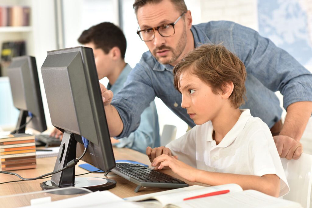 Student focused on typing at a computer while a teacher provides hands-on assistance.