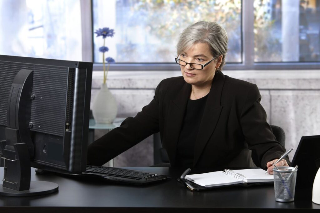Woman in an office environment taking notes while viewing information on a computer screen.