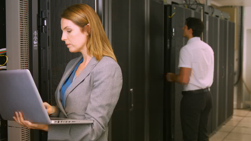 Female IT professional using a laptop while standing beside server racks in a data centre; another technician works in the background.