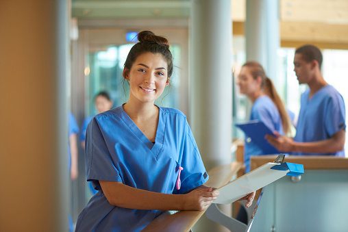 Young nurse standing with a clipboard while colleagues work in the background.