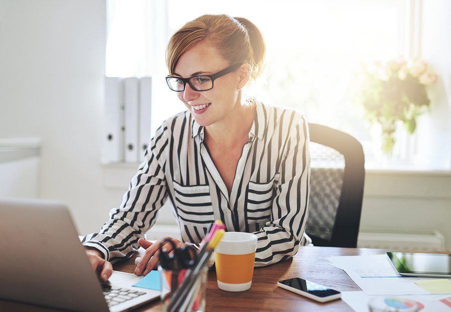 Smiling woman wearing glasses working on a laptop at her desk in a bright office.