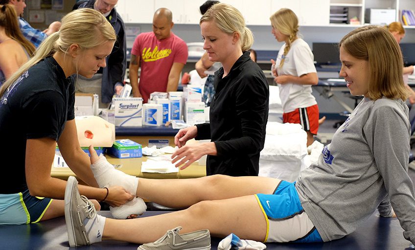 Therapist assisting patient with leg wrapping exercise in a clinical training lab.
