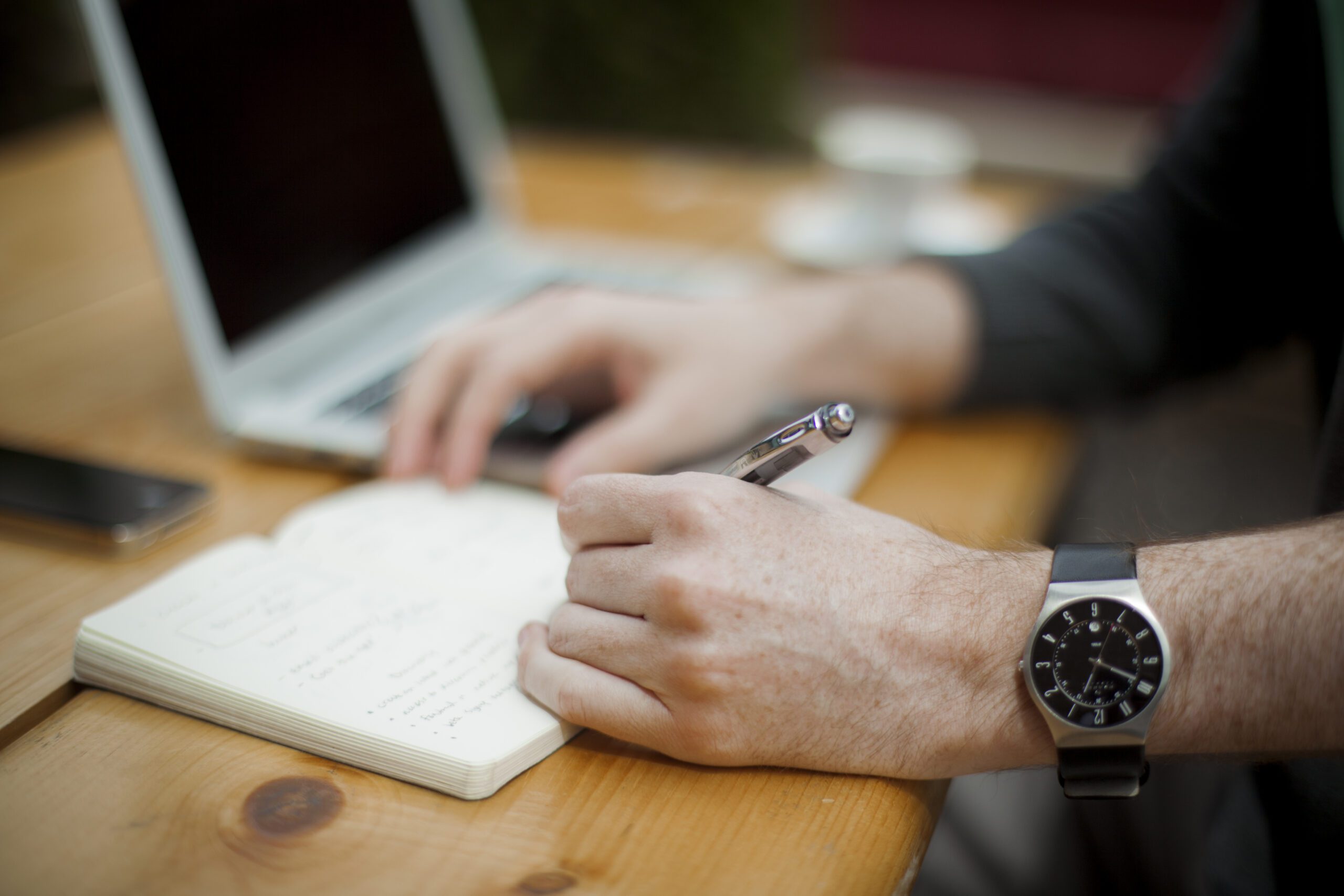 Person writing notes in a notebook while working on a laptop at a wooden desk.