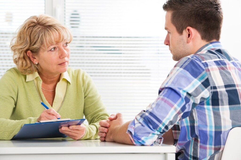 Healthcare professional speaking with a male patient while taking notes on a clipboard.