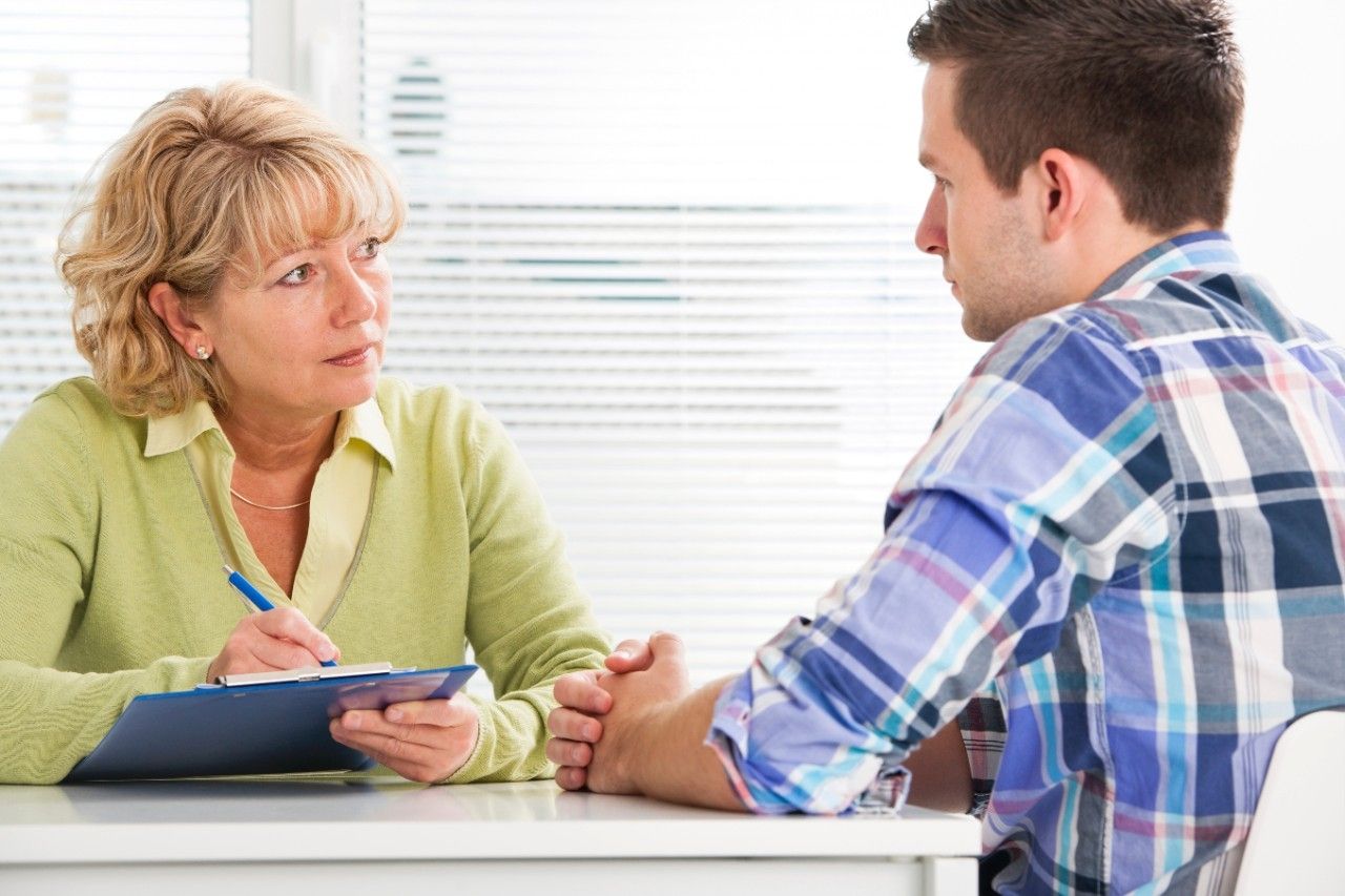 Healthcare professional speaking with a male patient while taking notes on a clipboard.