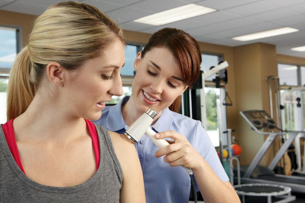 Physiotherapist using a handheld therapy device on a woman’s shoulder during a treatment session.