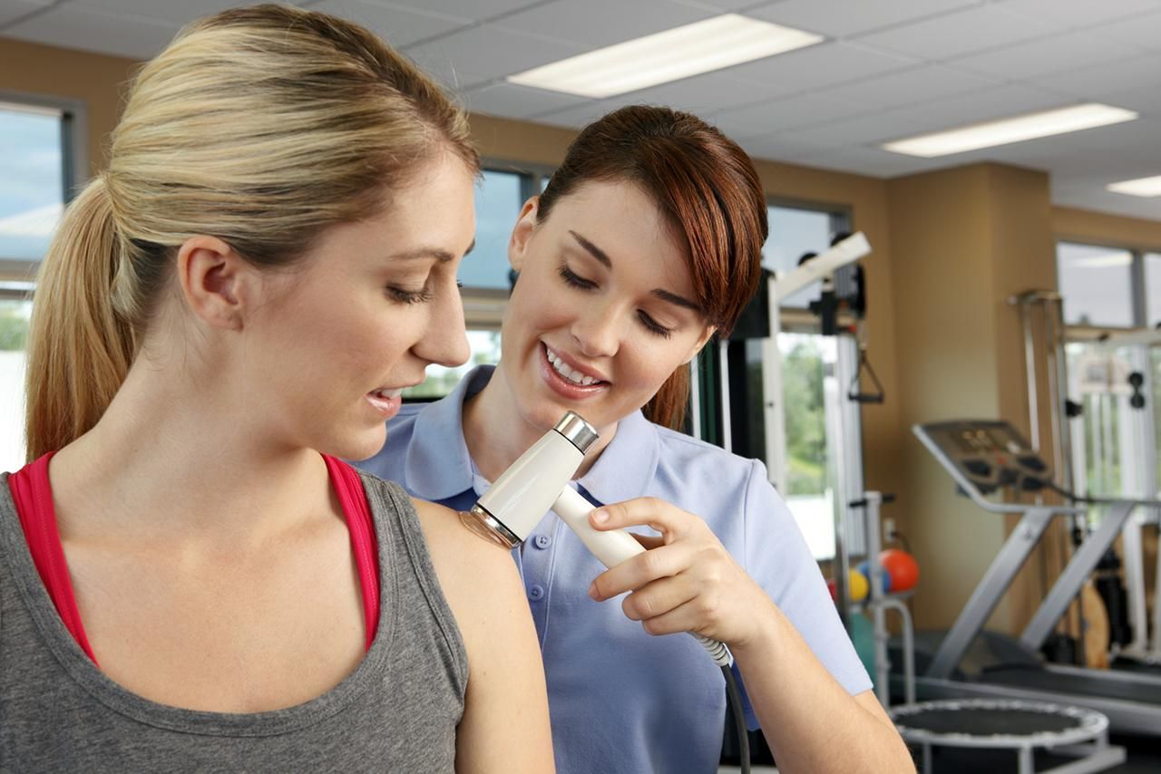 Physiotherapist using a handheld therapy device on a woman’s shoulder during a treatment session.