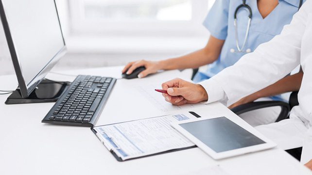 Doctors reviewing patient forms on a clipboard with a tablet and computer at a medical office desk.