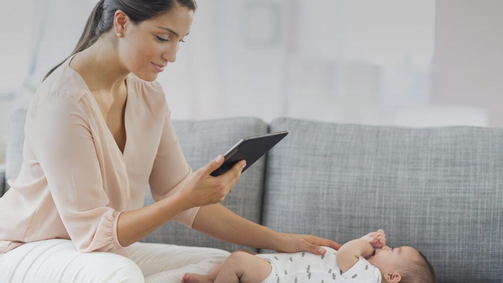 Mother sitting on a couch using a tablet while gently touching her baby lying beside her.