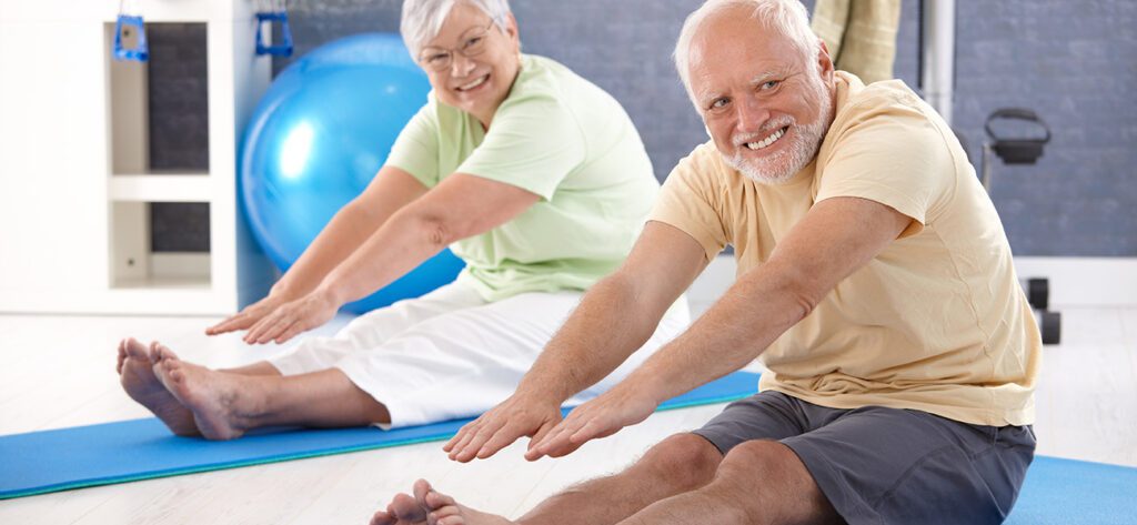 Smiling senior man and woman stretching forward on exercise mats during a fitness or physiotherapy session.