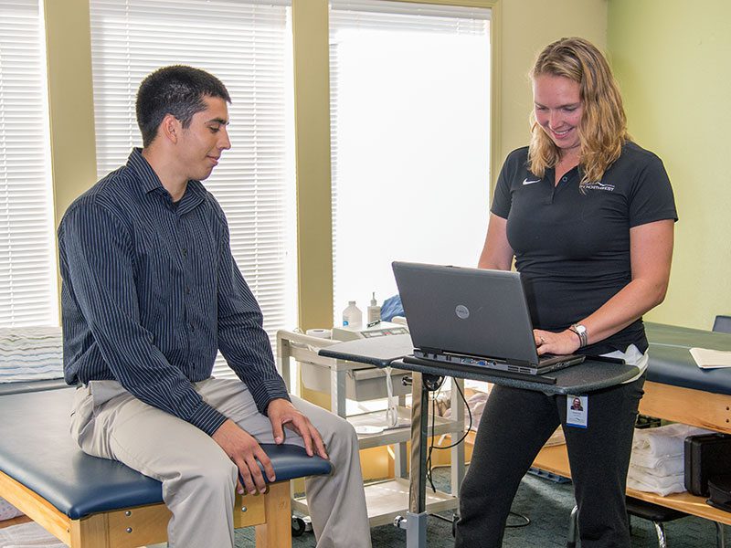 Physiotherapist entering notes on a laptop while discussing treatment with a patient seated on an exam table.
