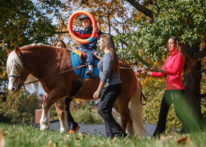 Child participating in equine therapy session with therapists guiding the horse outdoors.