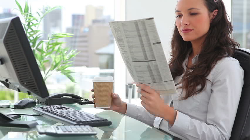 Professional woman reading newsletter at office desk with computer and coffee.