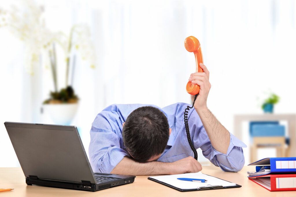 Frustrated office worker holding phone while resting head on desk beside laptop.