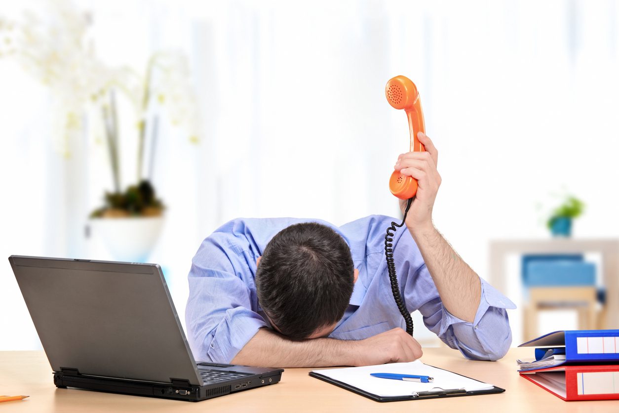 Frustrated office worker holding phone while resting head on desk beside laptop.