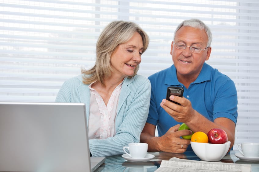 Happy mature couple reviewing information on smartphone and laptop at kitchen table.