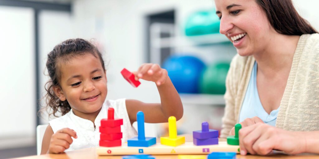Young girl developing fine motor skills with therapist using wooden stacking toy in a rehabilitation setting.