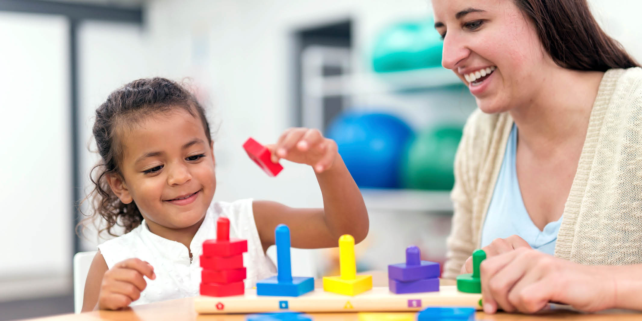 Young girl developing fine motor skills with therapist using wooden stacking toy in a rehabilitation setting.