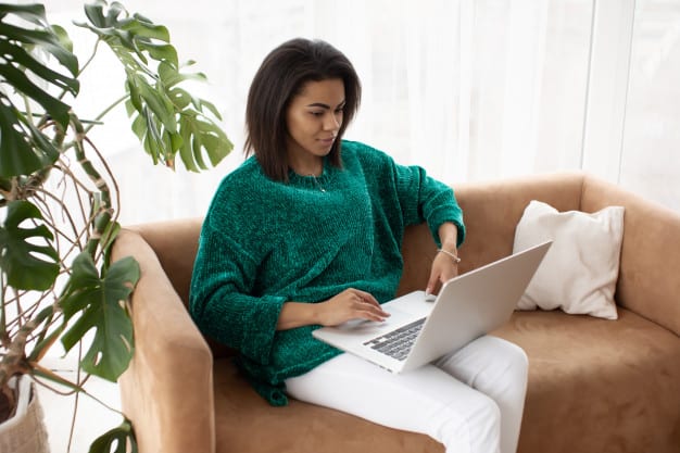 Woman sitting on a couch using laptop for remote work in bright modern living room.