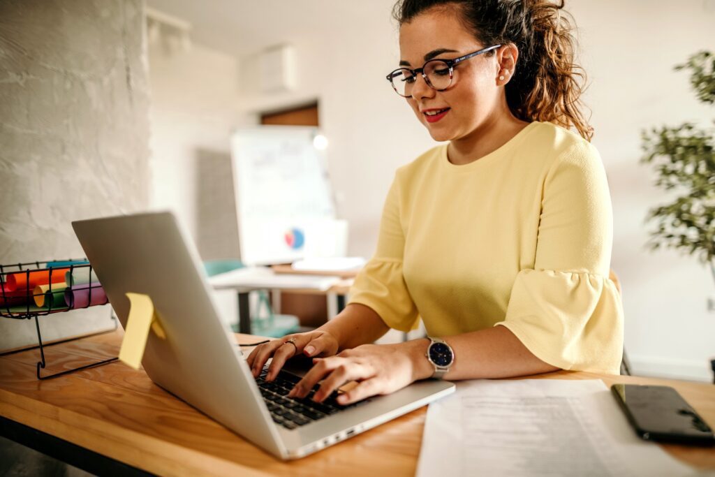 Businesswoman wearing glasses and a yellow blouse using a laptop at work.