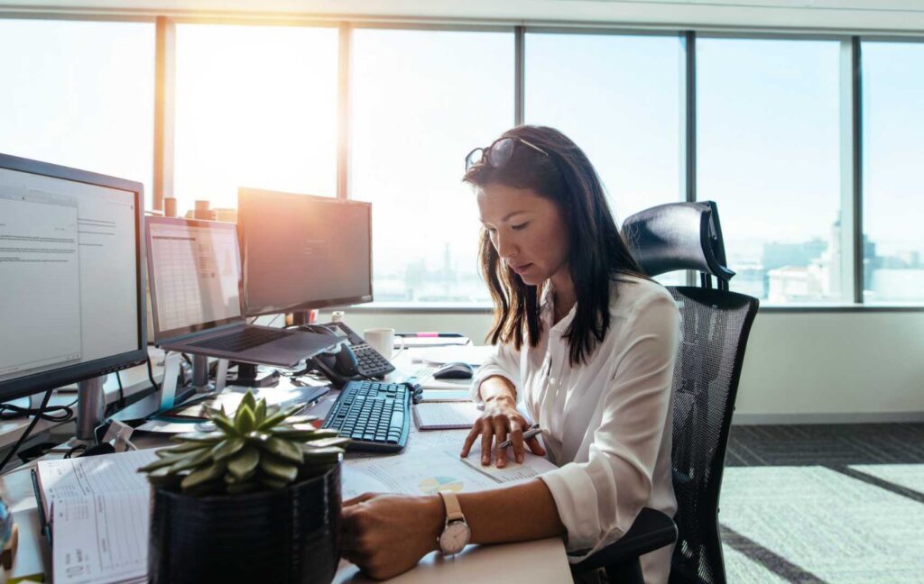 Business professional reviewing financial documents and data on multiple computer screens in a modern office.