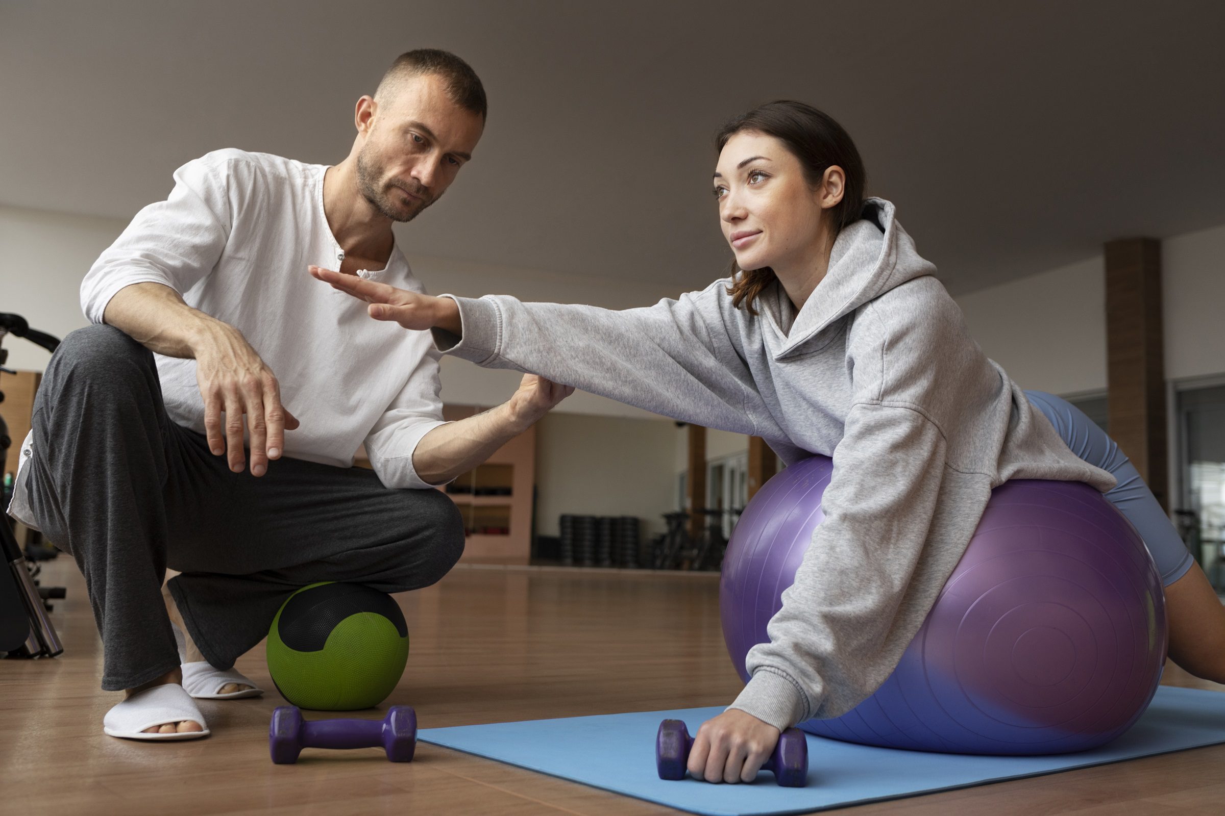 Physiotherapist assisting woman with balance exercise on stability ball during rehab session.