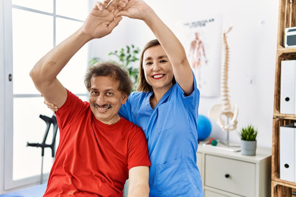 Smiling physiotherapist helping senior man stretch his arm during rehabilitation session.
