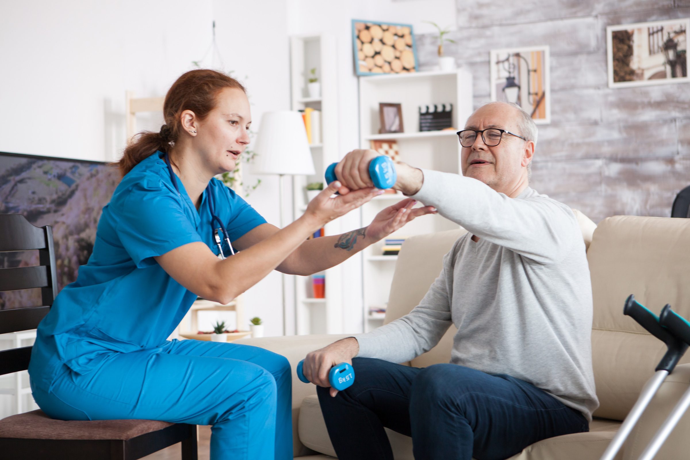 Therapist in blue scrubs helping elder gentleman lift light weight during rehabilitation session.
